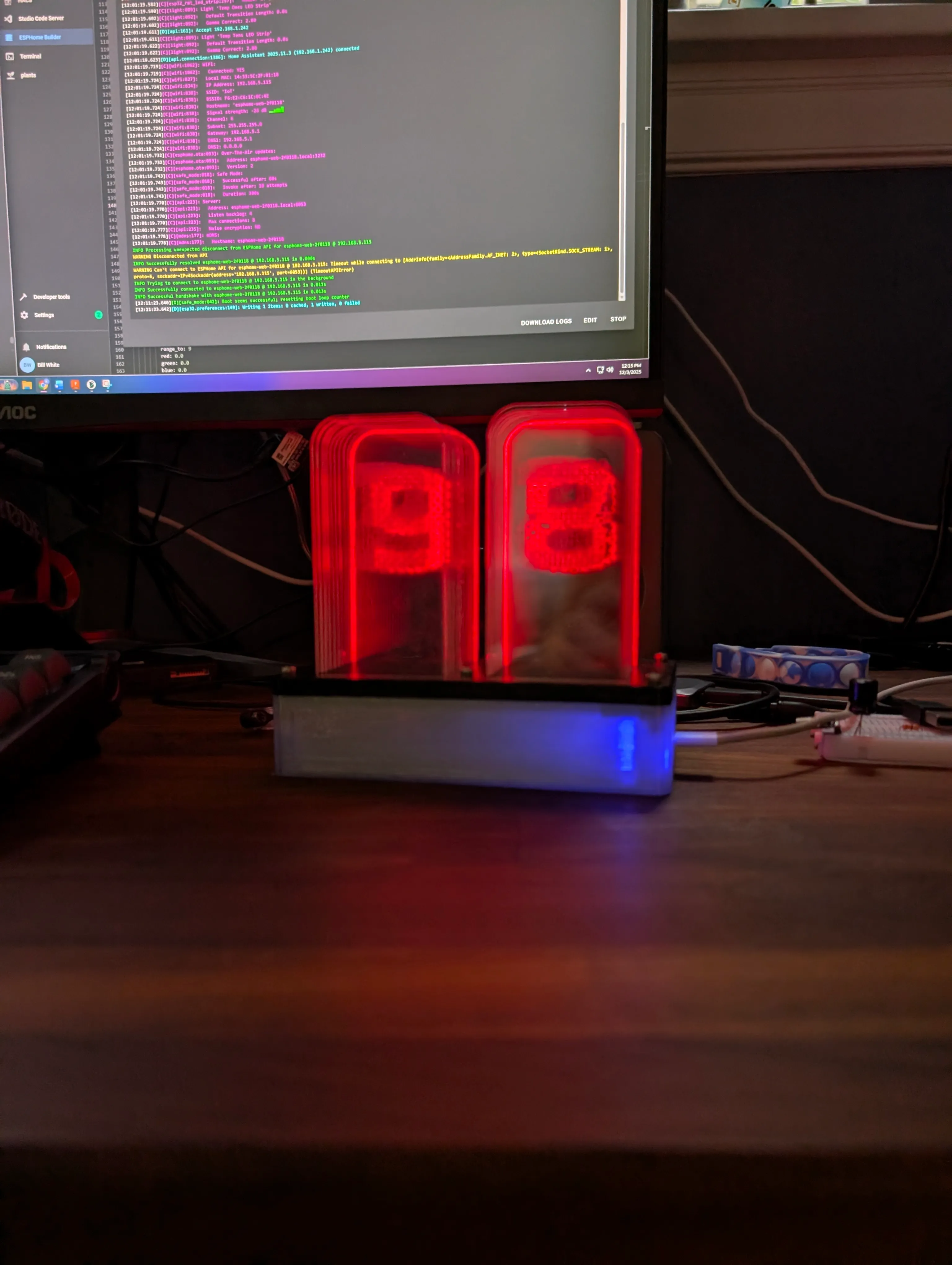 The thermometer lit in red, the digits reading 98, an ESPHome log open on the monitor behind it
